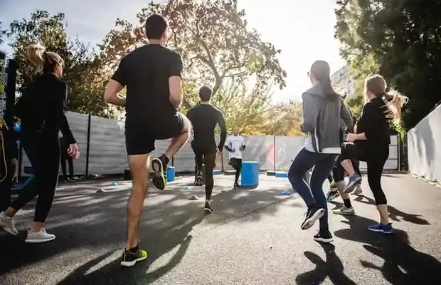 A group of individuals jogs together outdoors, with trees and sunlight in the background, engaged in a fitness workout.