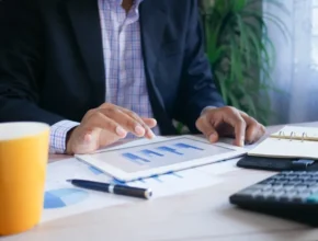 A professional man analyzes data on a tablet while seated at a desk with a coffee cup, notebook, and calculator.