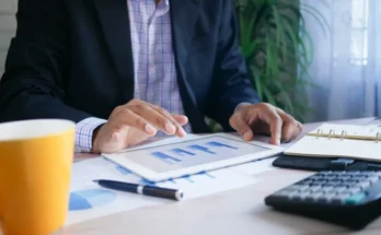 A professional man analyzes data on a tablet while seated at a desk with a coffee cup, notebook, and calculator.