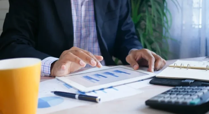 A professional man analyzes data on a tablet while seated at a desk with a coffee cup, notebook, and calculator.