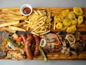 A wooden platter featuring assorted grilled meats, sausages, fries, fried plantains, and a small bowl of sauce.