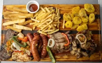 A wooden platter featuring assorted grilled meats, sausages, fries, fried plantains, and a small bowl of sauce.