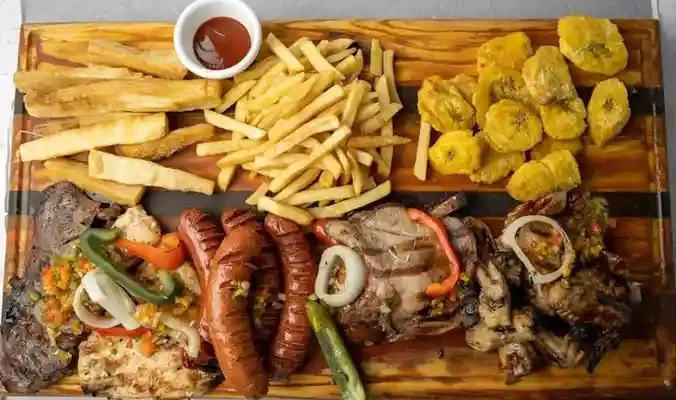 A wooden platter featuring assorted grilled meats, sausages, fries, fried plantains, and a small bowl of sauce.