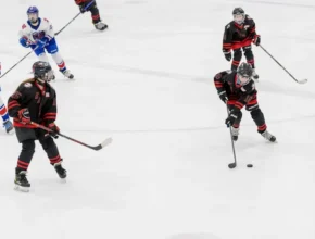 Youth ice hockey players in black and red jerseys compete on the rink, focused on the puck as they maneuver and strategize.