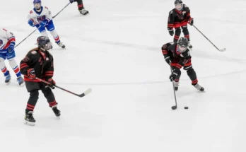 Youth ice hockey players in black and red jerseys compete on the rink, focused on the puck as they maneuver and strategize.