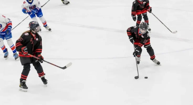 Youth ice hockey players in black and red jerseys compete on the rink, focused on the puck as they maneuver and strategize.