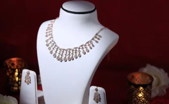 Elegant rose gold necklace displayed on a bust, accompanied by matching earrings, set against a festive red backdrop with candles.