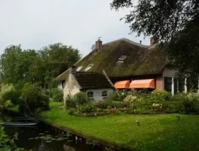 A charming thatched-roof house with orange awnings, surrounded by a lush garden and a serene canal, under a cloudy sky.