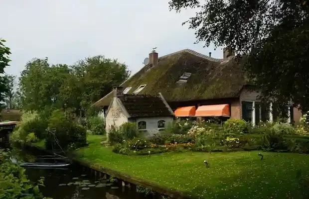 A charming thatched-roof house with orange awnings, surrounded by a lush garden and a serene canal, under a cloudy sky.