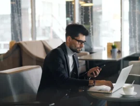 A man in a stylish outfit uses a laptop and checks his phone while sitting at a café table, with a coffee cup beside him.