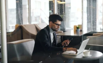 A man in a stylish outfit uses a laptop and checks his phone while sitting at a café table, with a coffee cup beside him.