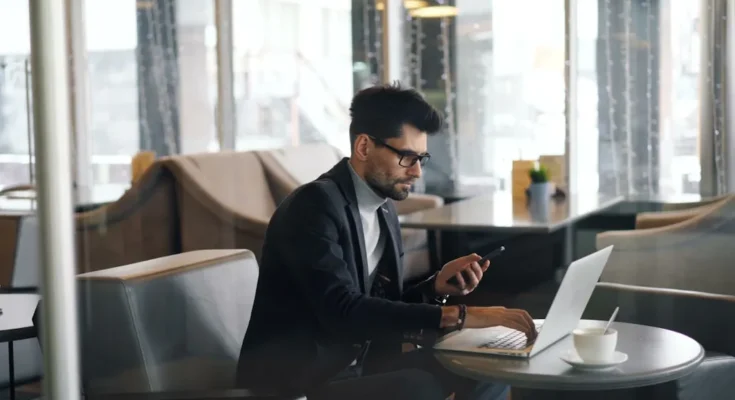 A man in a stylish outfit uses a laptop and checks his phone while sitting at a café table, with a coffee cup beside him.
