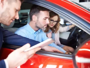 A couple sits inside a red car at a dealership, engaging with a salesperson holding a tablet outside the vehicle.