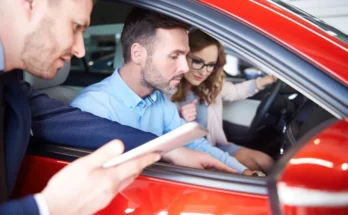 A couple sits inside a red car at a dealership, engaging with a salesperson holding a tablet outside the vehicle.