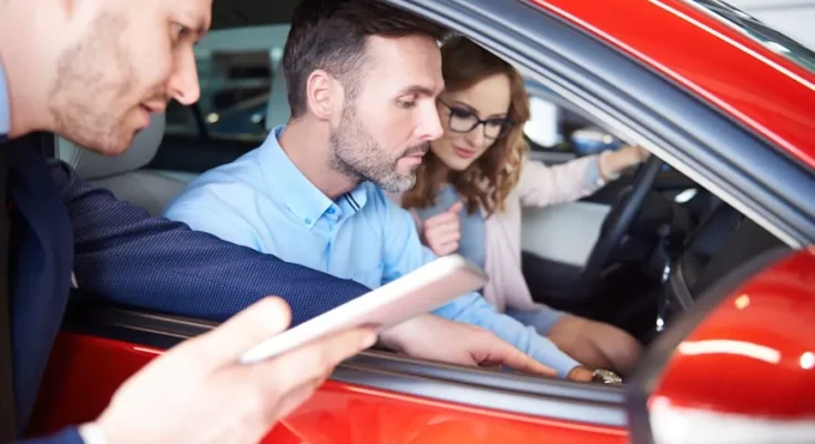 A couple sits inside a red car at a dealership, engaging with a salesperson holding a tablet outside the vehicle.