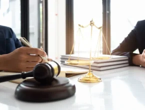 A close-up of a lawyer writing notes beside a gavel and a golden scale of justice in a well-lit office setting.
