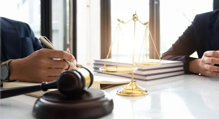 A close-up of a lawyer writing notes beside a gavel and a golden scale of justice in a well-lit office setting.