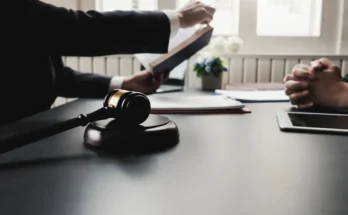 A lawyer presents a book while a client listens attentively; a gavel sits on the table, emphasizing a legal discussion.