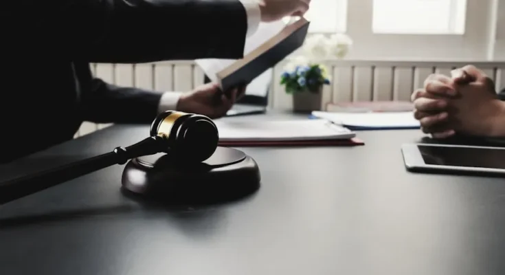 A lawyer presents a book while a client listens attentively; a gavel sits on the table, emphasizing a legal discussion.