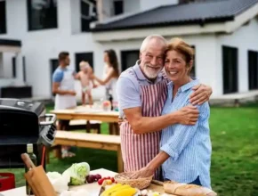 A happy couple embrace at a sunny outdoor gathering, surrounded by fresh vegetables and a barbecue, while the family enjoys the moment.