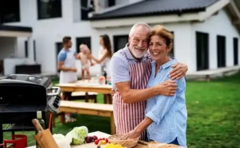 A happy couple embrace at a sunny outdoor gathering, surrounded by fresh vegetables and a barbecue, while the family enjoys the moment.