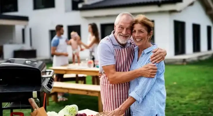 A happy couple embrace at a sunny outdoor gathering, surrounded by fresh vegetables and a barbecue, while the family enjoys the moment.