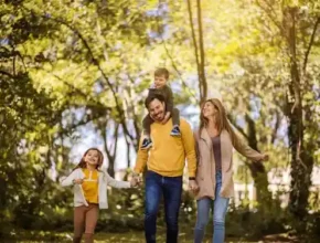 A family joyfully walks hand in hand along a sunlit path, surrounded by lush greenery and golden autumn leaves.