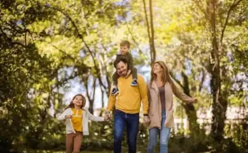 A family joyfully walks hand in hand along a sunlit path, surrounded by lush greenery and golden autumn leaves.