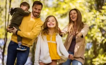 A joyful family enjoys a day outdoors, with two adults and two children playing among vibrant greenery in the background.