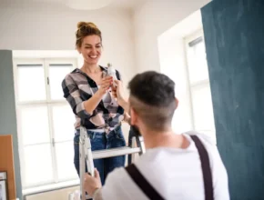 A woman stands on a ladder, holding a paint roller, while a man assists her from below in a brightly lit room.