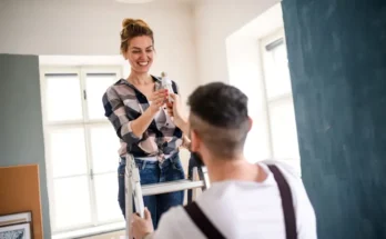 A woman stands on a ladder, holding a paint roller, while a man assists her from below in a brightly lit room.