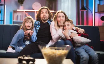A family of four sits closely together on a couch, smiling and enjoying time together, with a bowl of popcorn in the foreground.