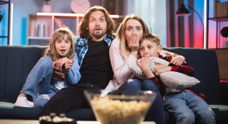 A family of four sits closely together on a couch, smiling and enjoying time together, with a bowl of popcorn in the foreground.