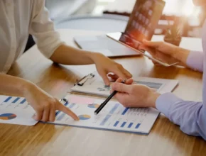 Two people collaboratively reviewing financial charts and reports on a table, with one pointing and the other taking notes.