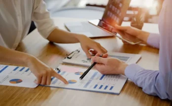 Two people collaboratively reviewing financial charts and reports on a table, with one pointing and the other taking notes.