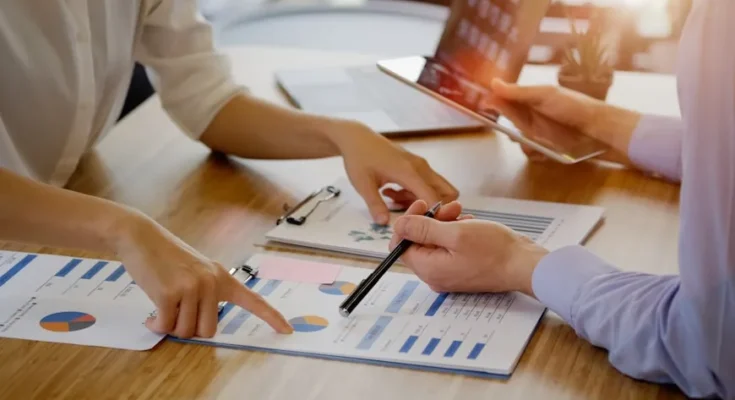 Two people collaboratively reviewing financial charts and reports on a table, with one pointing and the other taking notes.