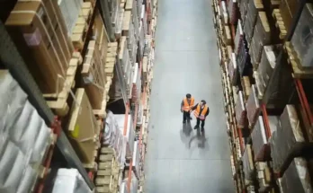 Two warehouse workers in orange vests walk between tall shelves stacked with boxes, observing the organized storage space.