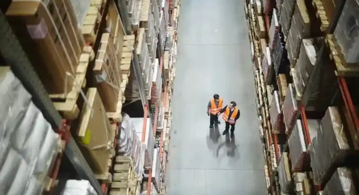 Two warehouse workers in orange vests walk between tall shelves stacked with boxes, observing the organized storage space.