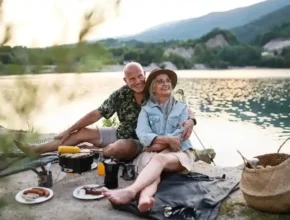A couple enjoys a picnic by a lake at sunset, surrounded by nature, with plates of food and cooking equipment nearby.