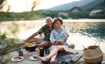 A couple enjoys a picnic by a lake at sunset, surrounded by nature, with plates of food and cooking equipment nearby.