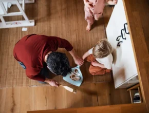 A parent serves snacks to a child seated on the floor, while another child watches nearby in a cozy home setting.