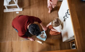 A parent serves snacks to a child seated on the floor, while another child watches nearby in a cozy home setting.