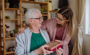 An older man receives a wrapped gift box from a younger woman indoors; both are smiling and wearing glasses.