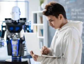 A person in a white hoodie types on a laptop, while a futuristic robot stands nearby in a modern workspace.