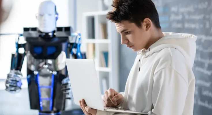A person in a white hoodie types on a laptop, while a futuristic robot stands nearby in a modern workspace.