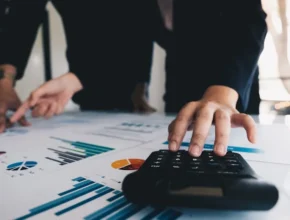 Two business professionals analyze charts and graphs on papers while one operates a calculator on a desk.