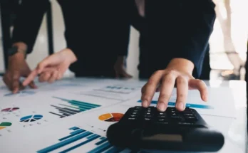 Two business professionals analyze charts and graphs on papers while one operates a calculator on a desk.