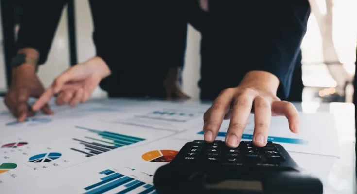 Two business professionals analyze charts and graphs on papers while one operates a calculator on a desk.