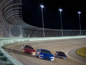 Three race cars, one red, one blue, and one black, compete closely on a lit oval track at night.