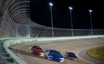 Three race cars, one red, one blue, and one black, compete closely on a lit oval track at night.